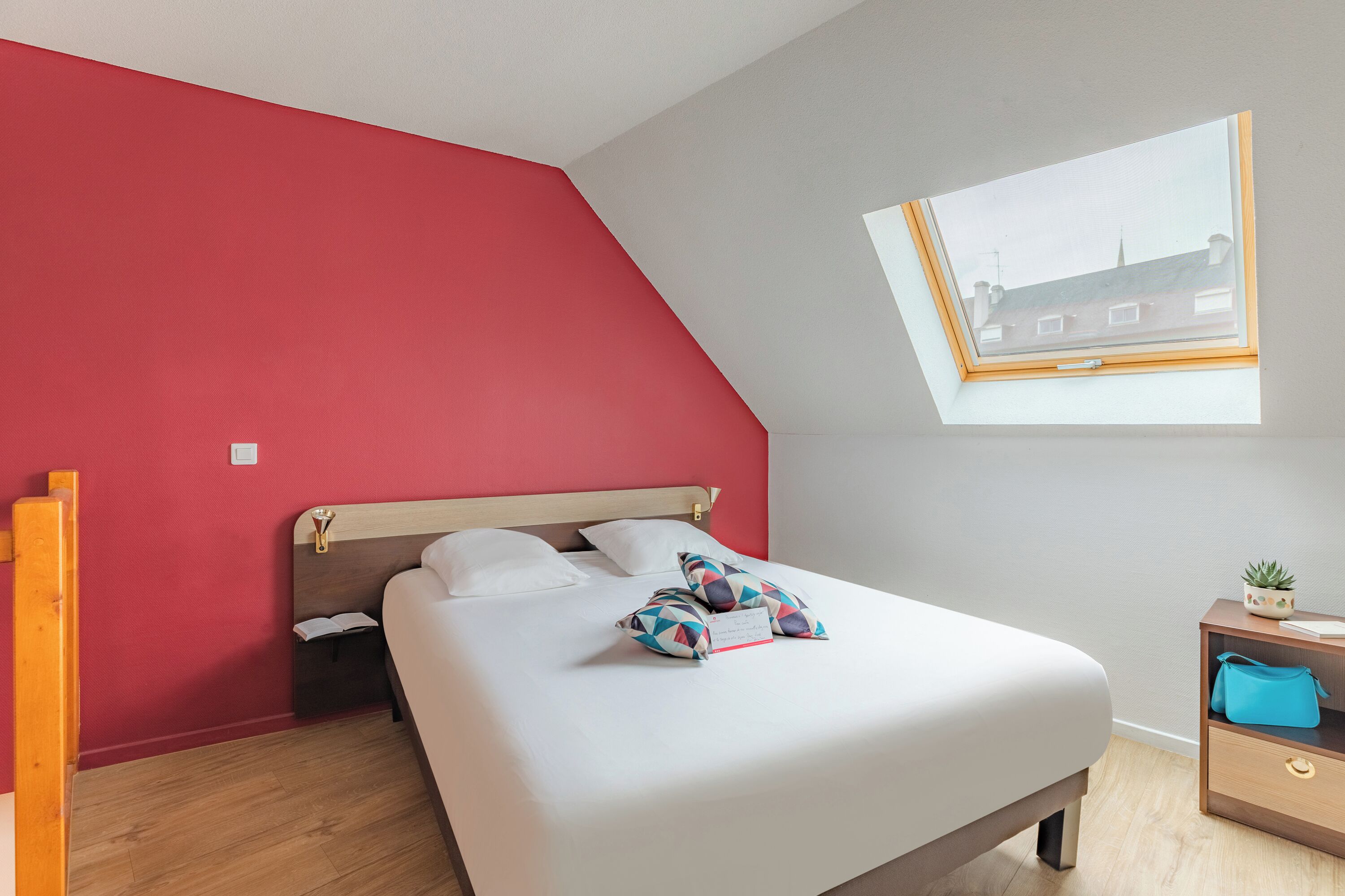 A bedroom with a sloped ceiling, a skylight, a bed with white bedding, a nightstand with a blue bag, and a small plant.