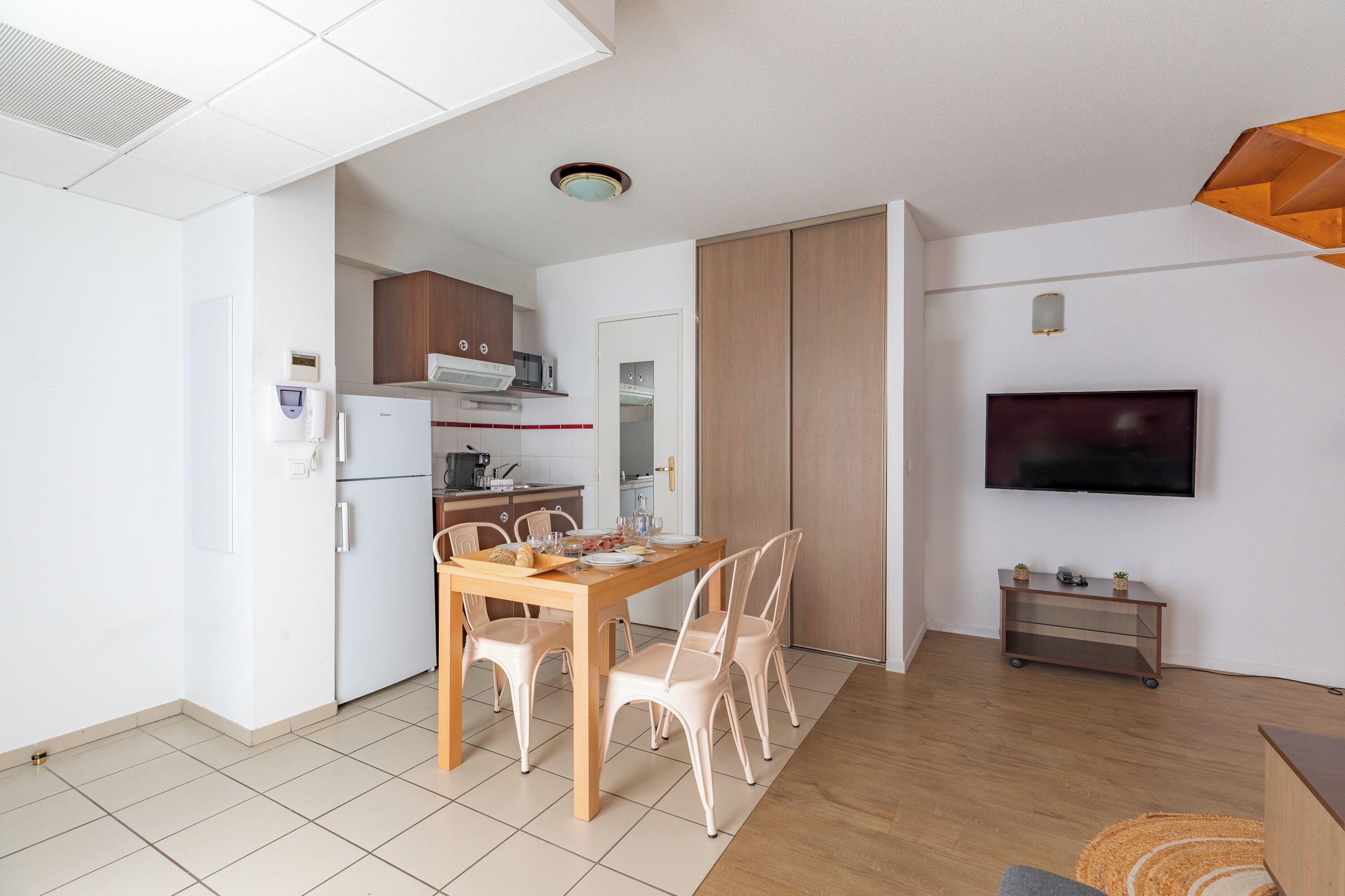 A modern kitchen with white cabinets, a dining table with chairs, a TV mounted on the wall, and a small cart with plants.