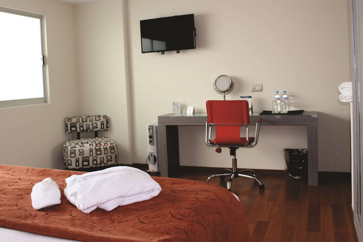 Une chambre d’hôtel avec un lit, un bureau avec un ordinateur, une chaise rouge, une télévision et une fenêtre.