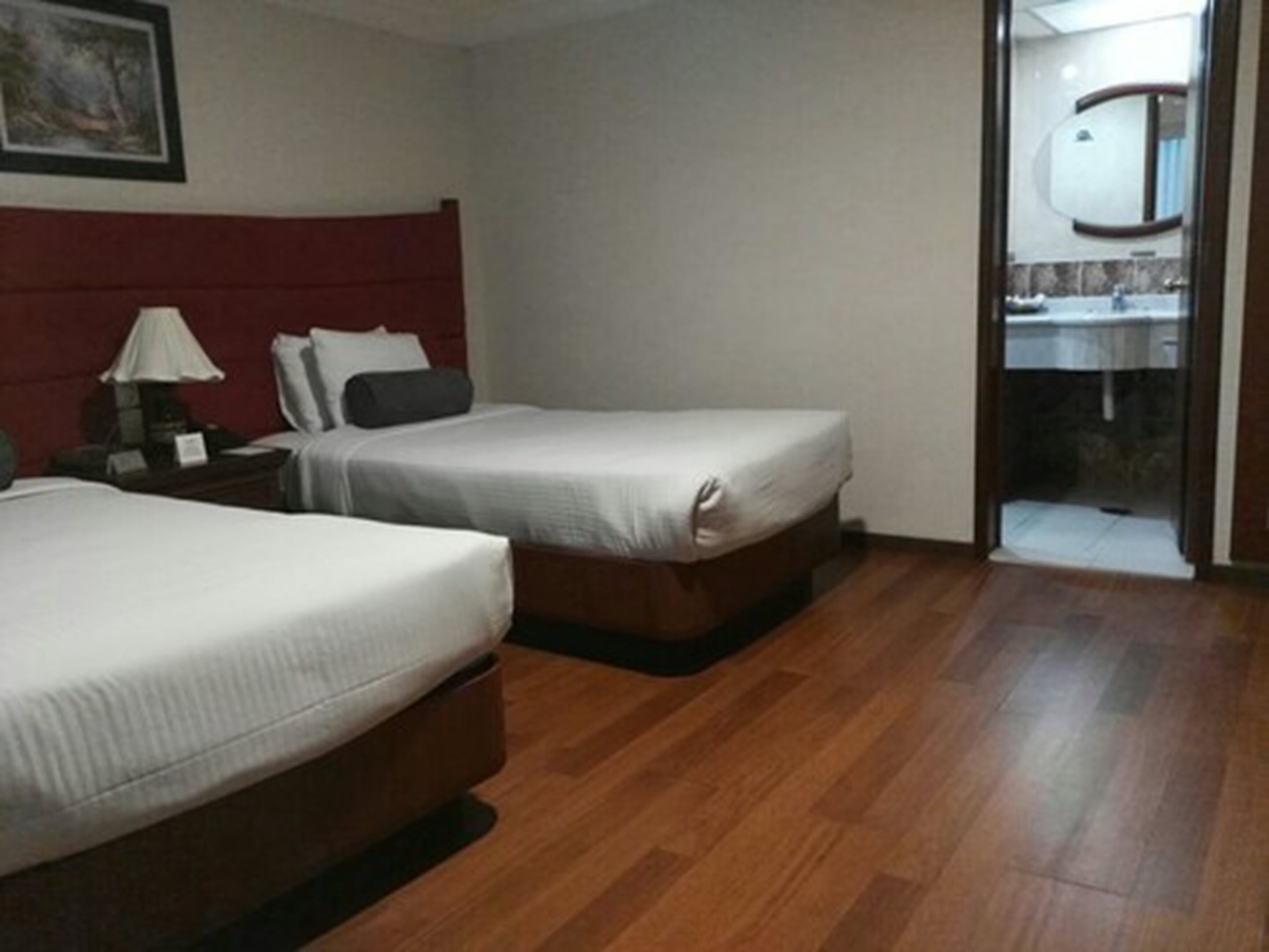 Camera d'albergo con due letti, pavimento in legno, testiera rossa, comodino con lampada e bagno visibile attraverso una porta aperta.