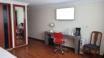 Habitación de hotel con piso de madera, un escritorio con computadora, una silla de oficina roja, una silla de madera, una cafetera y un televisor en la pared.