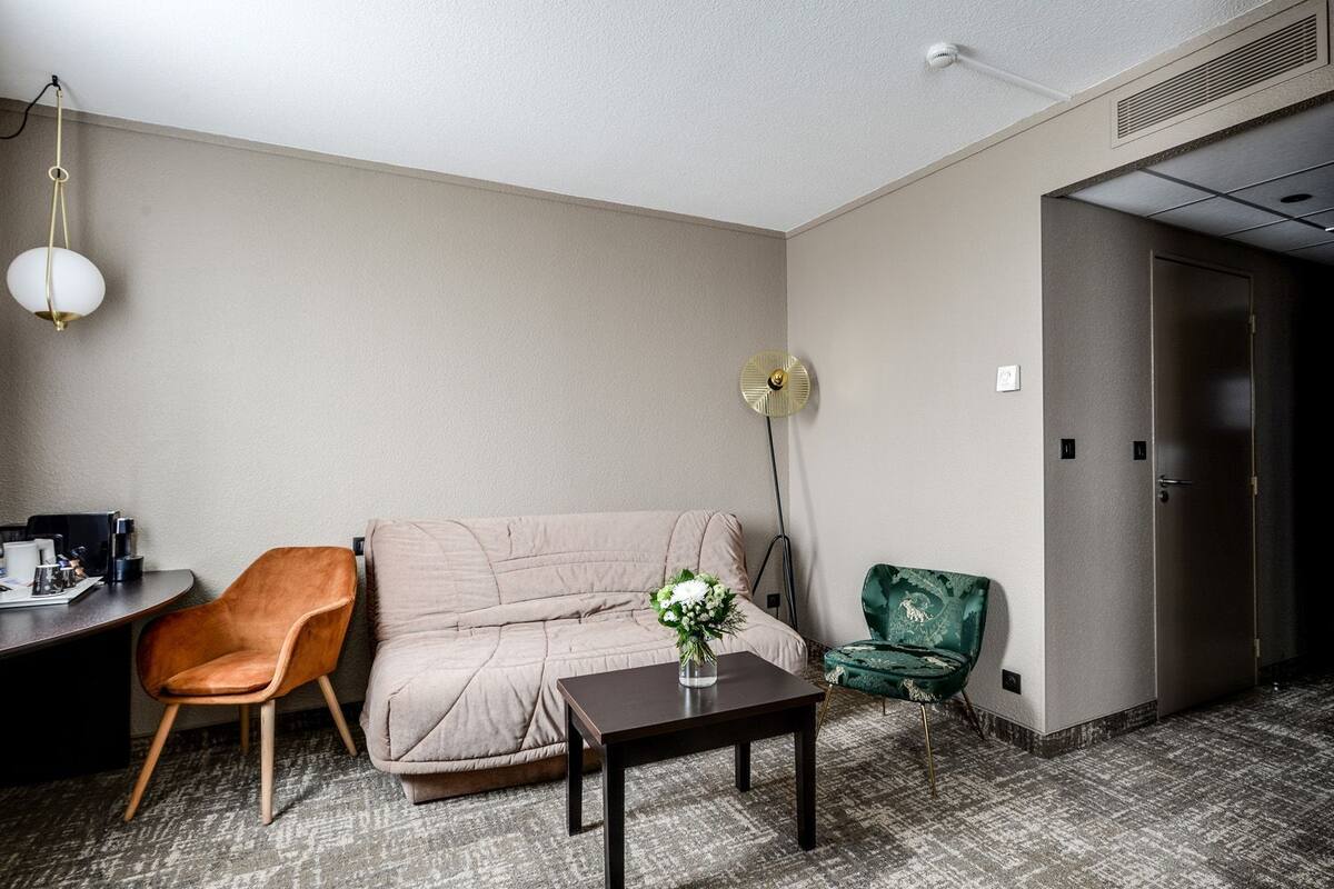 A hotel room with a sofa, a chair, a small table with a vase, a desk with a coffee machine, and a door.