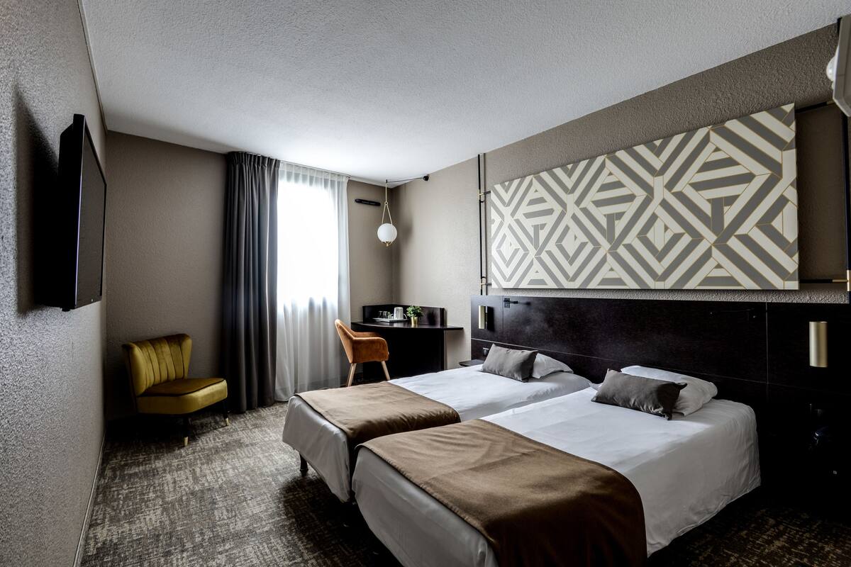 A hotel room with two beds, a desk, a chair, a TV, and a patterned wall.
