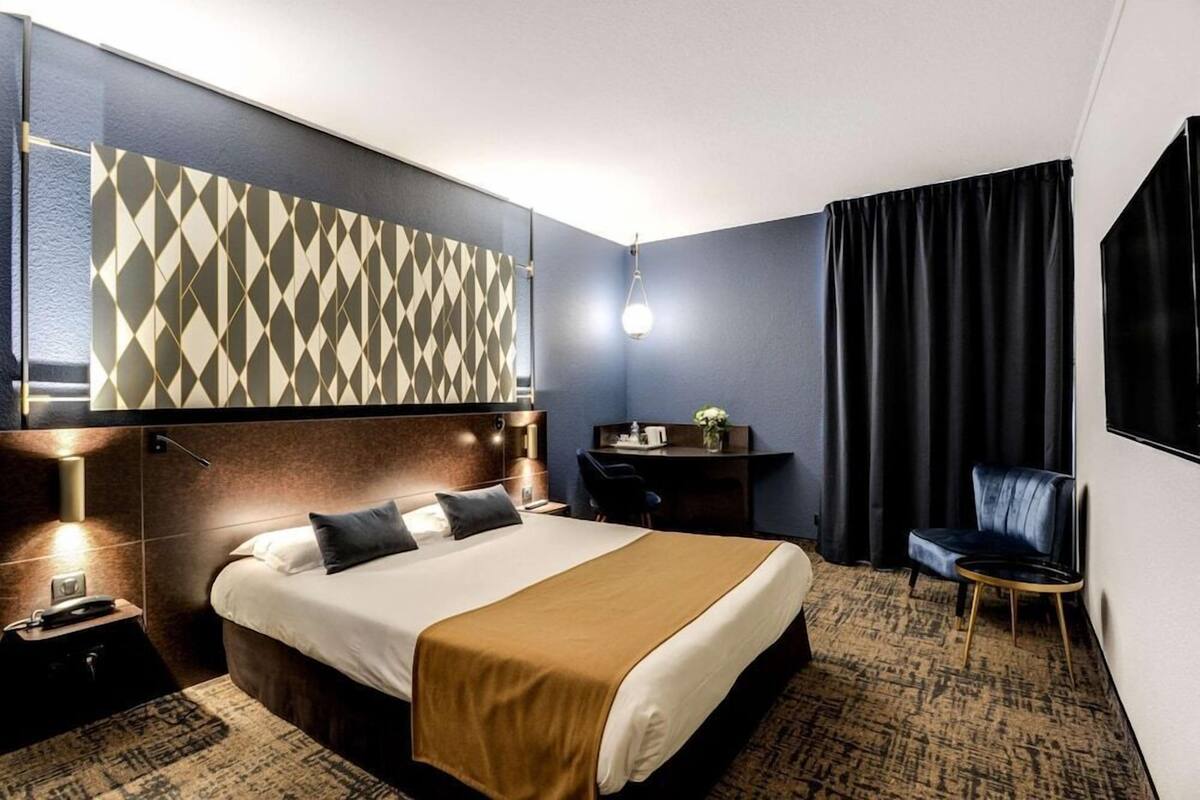 A hotel room with a bed, a desk, a chair, a TV, and a patterned wall.
