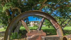 A large, rusted metal wheel structure in a park with a house in the background. - Habitation du Comté (Sainte-Rose)