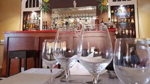 A bar area with two empty wine glasses on a table, shelves with various liquor bottles, and a mural with birds and foliage. - Habitation du Comté (Sainte-Rose)