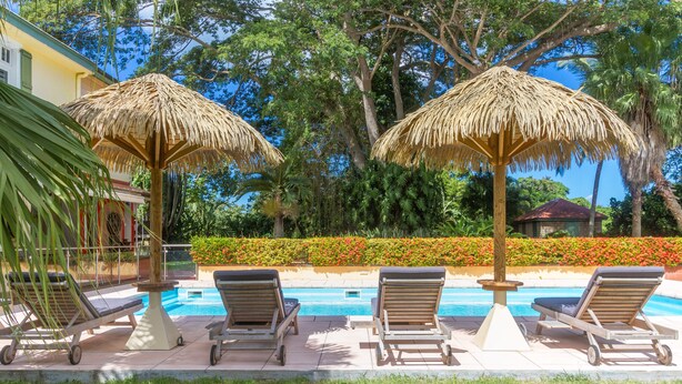 A pool area with lounge chairs and thatched umbrellas, surrounded by lush greenery and a clear blue sky. - Habitation du Comté (Sainte-Rose)