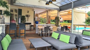 An outdoor seating area with grey sofas and tables, a bar, and a pool in the background. - Habitation du Comté (Sainte-Rose)