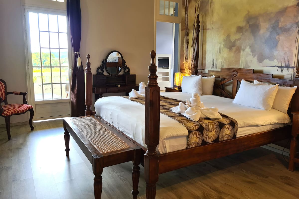 Une chambre bien éclairée, avec un grand lit, une table de chevet en bois, une commode, une chaise et une grande fenêtre.