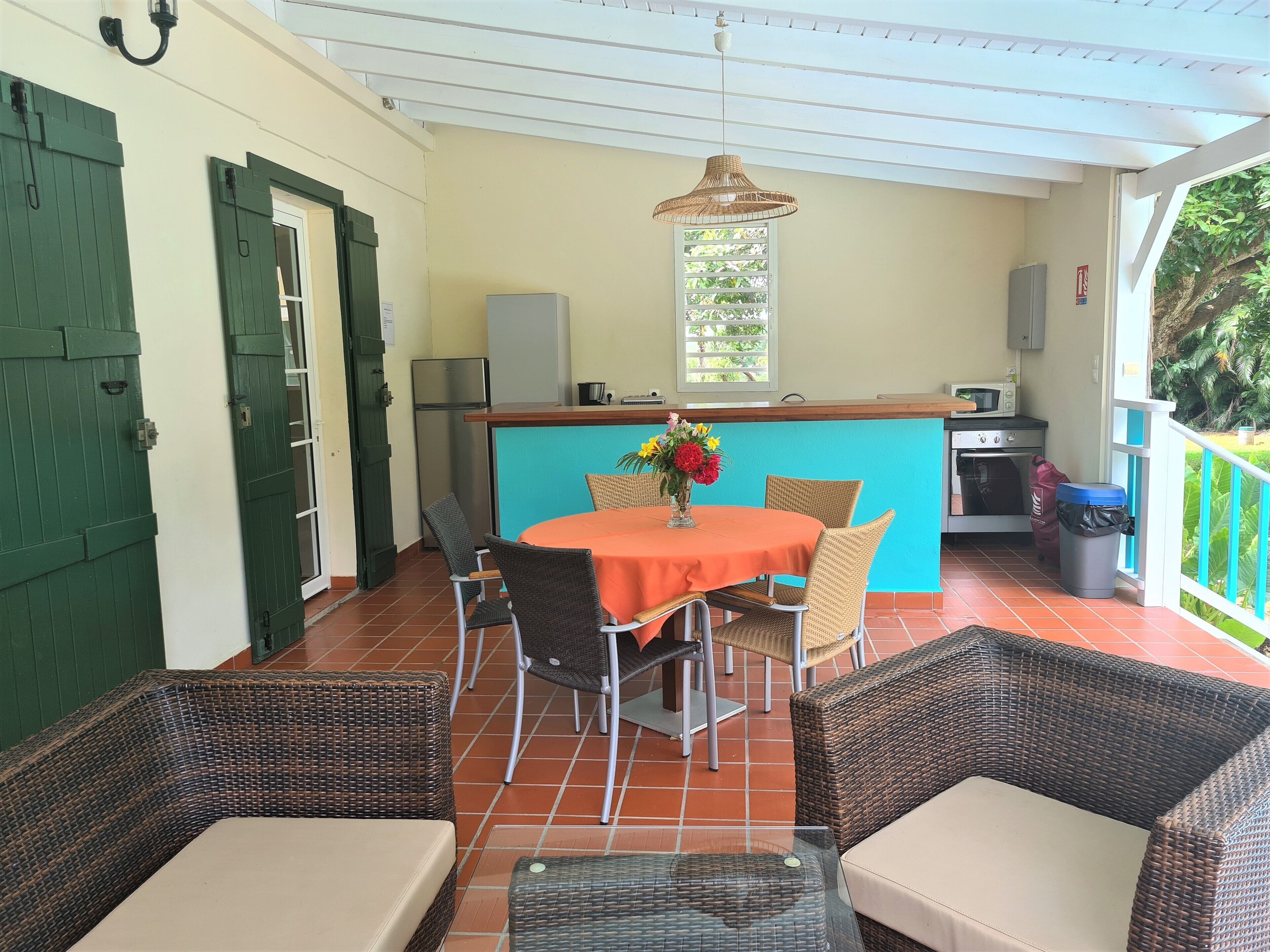 A covered patio area with wicker furniture, a central table with a vase of flowers, and a kitchen area in the background.