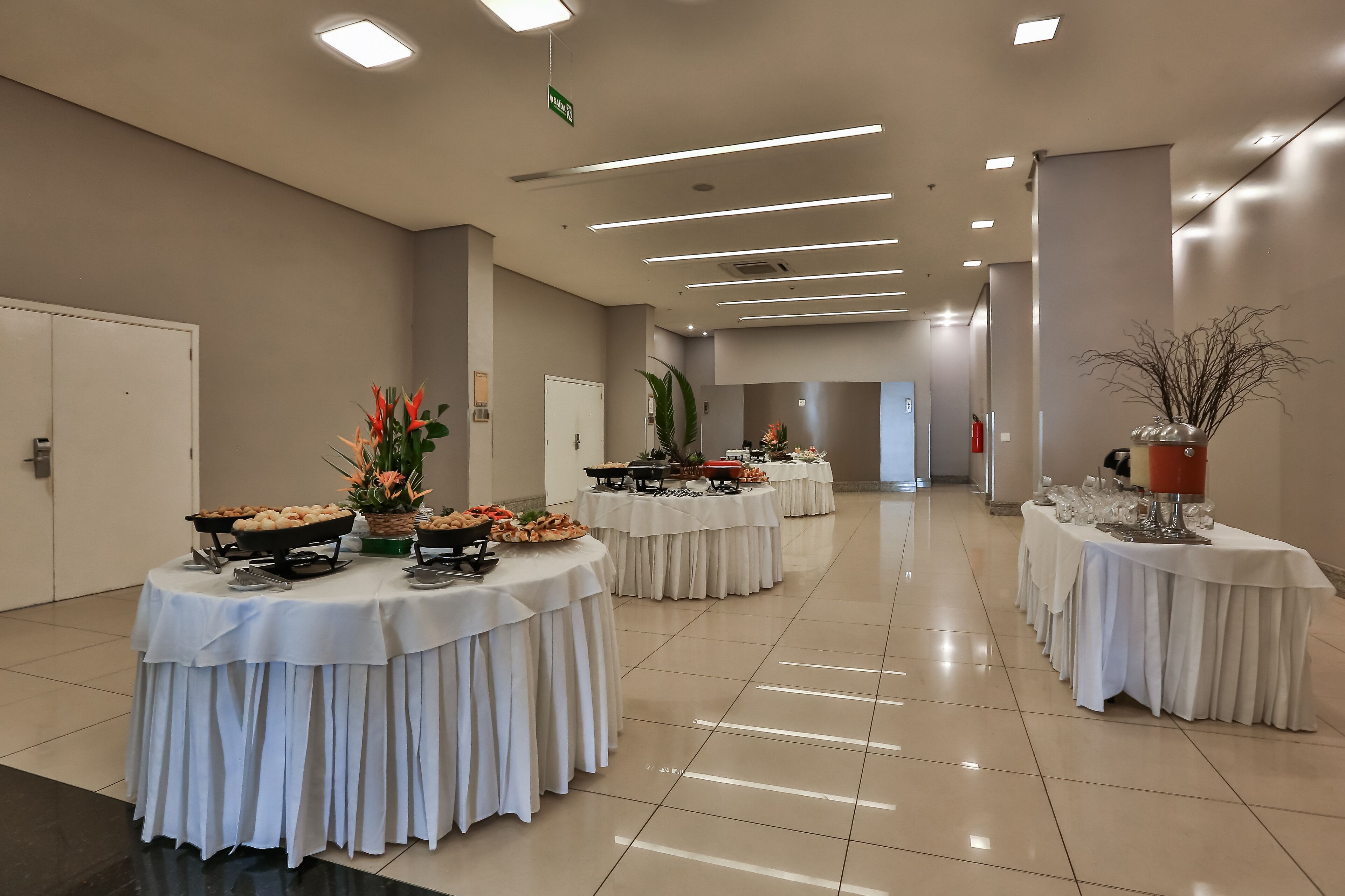 A spacious, well-lit conference room with multiple round tables covered in white tablecloths, each set with various food items and drinks.