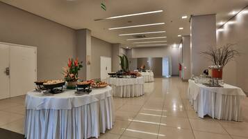A spacious, well-lit conference room with multiple round tables covered in white tablecloths, each set with various food items and drinks.