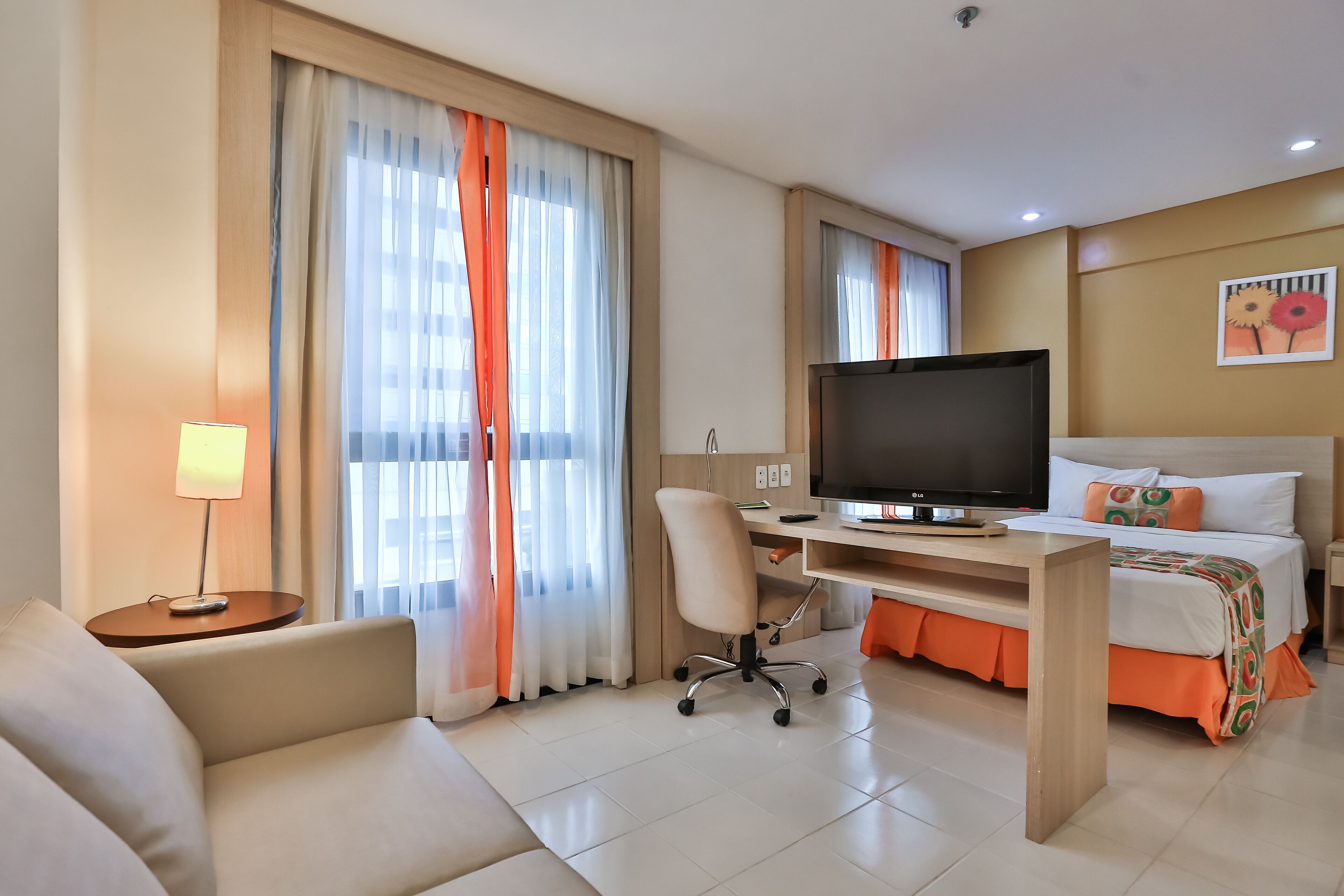 A hotel room with a bed, a desk with a TV, a chair, and a lamp.