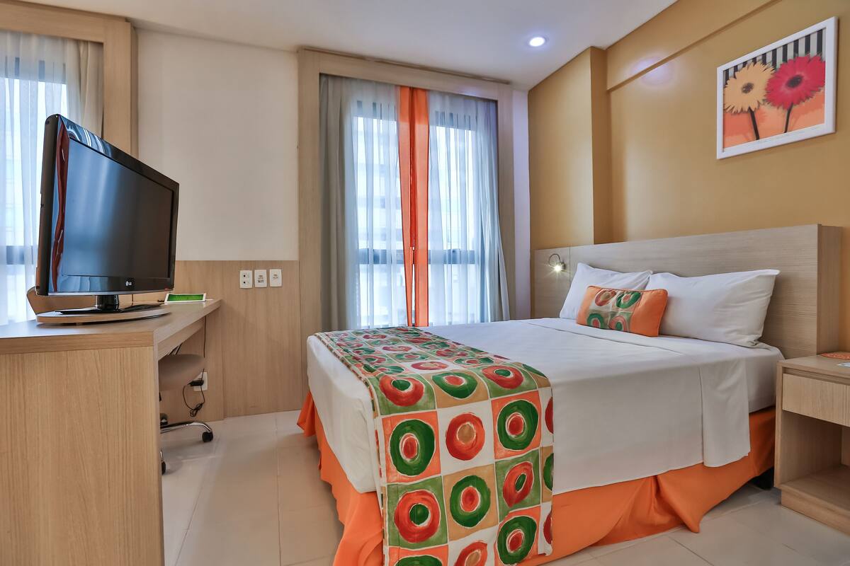 A hotel room with a bed, a desk with a television, and a window with curtains.