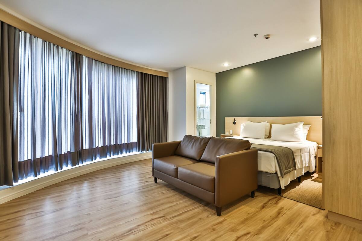 A modern hotel room with a wooden floor, a brown leather sofa, a bed with white bedding, and a large window with curtains.