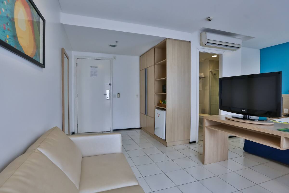A modern living room with a beige sofa, a wooden desk with a flat-screen TV, a small white fridge, and a built-in shelving unit.