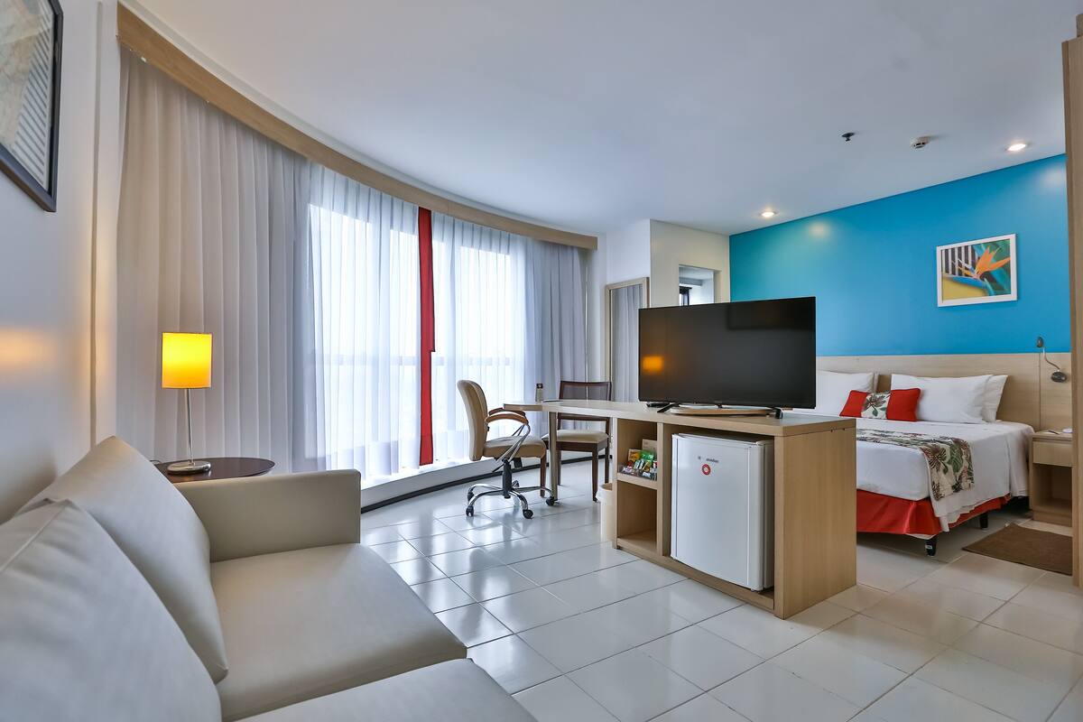 A hotel room with a sofa, a desk with a TV, a bed, and a large window with curtains.