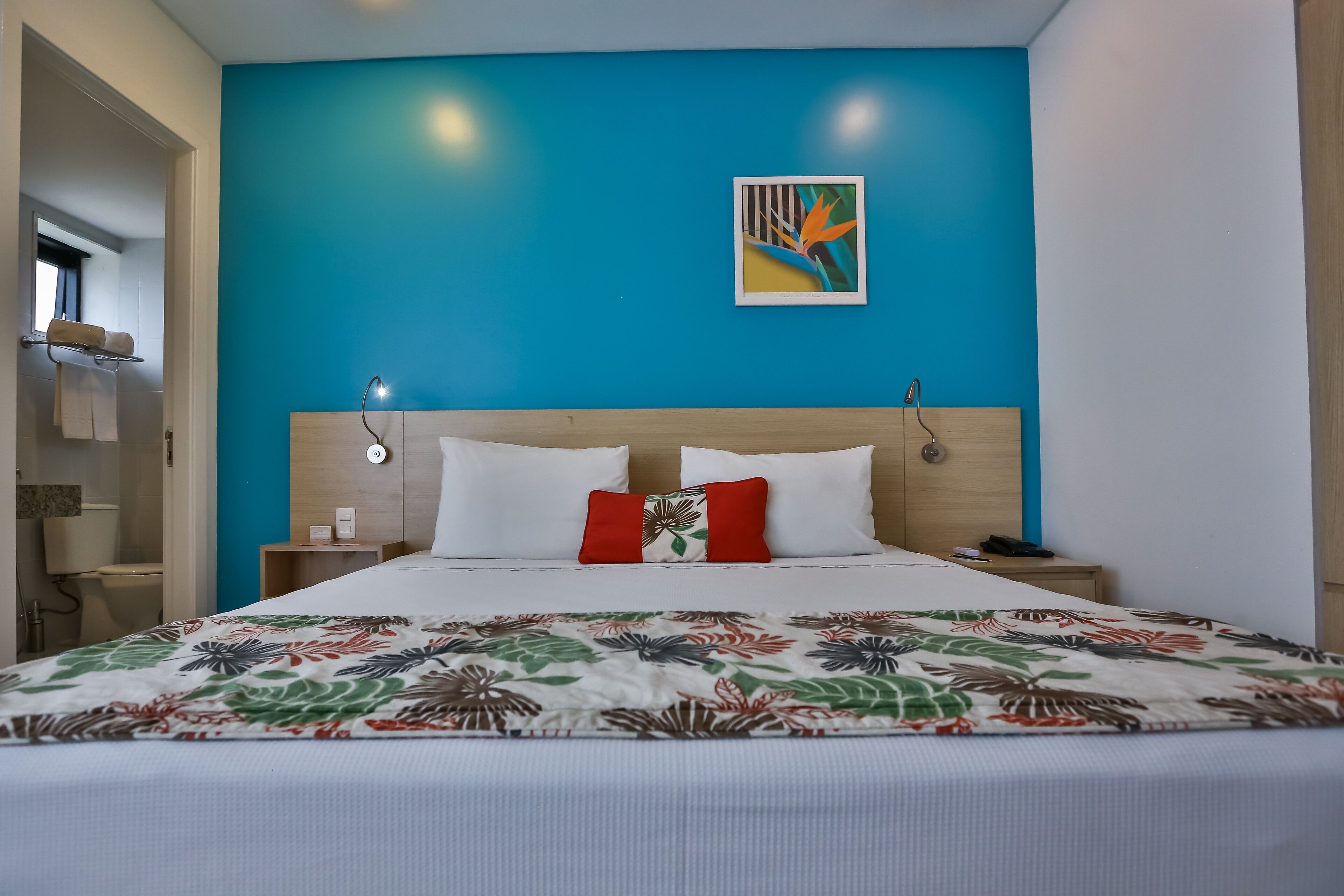 Quarto de hotel com cama, parede azul decorativa, um quadro e cabeceira de madeira.