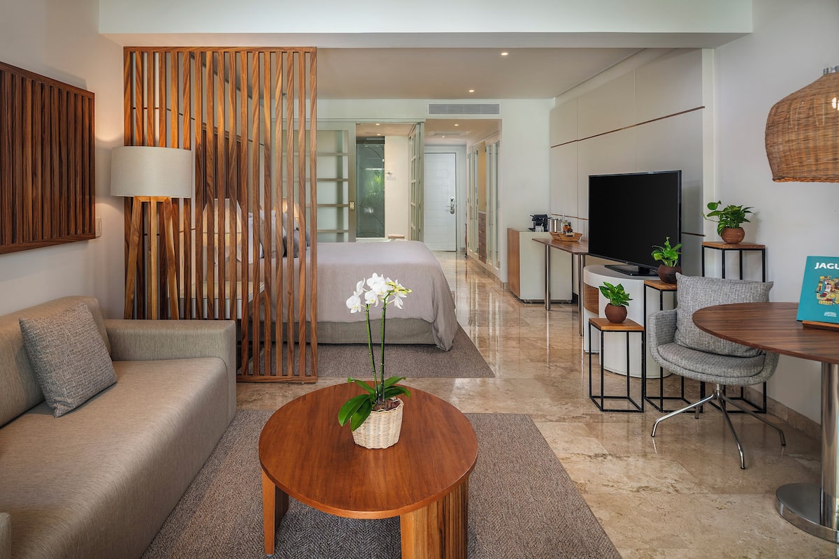 A modern hotel room with a sofa, a round wooden table with a plant, a TV, and a dining area with a chair.