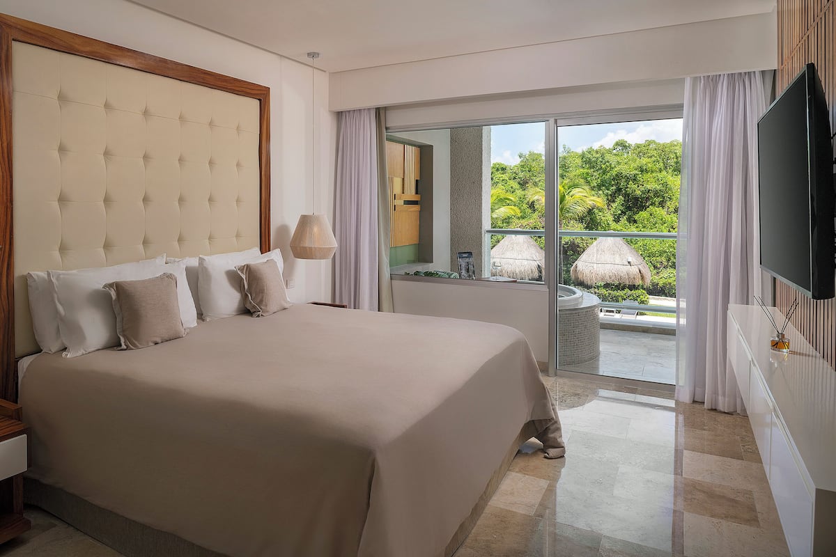 A large bed with a tufted headboard, a television, and a view of a lush garden through a sliding glass door.