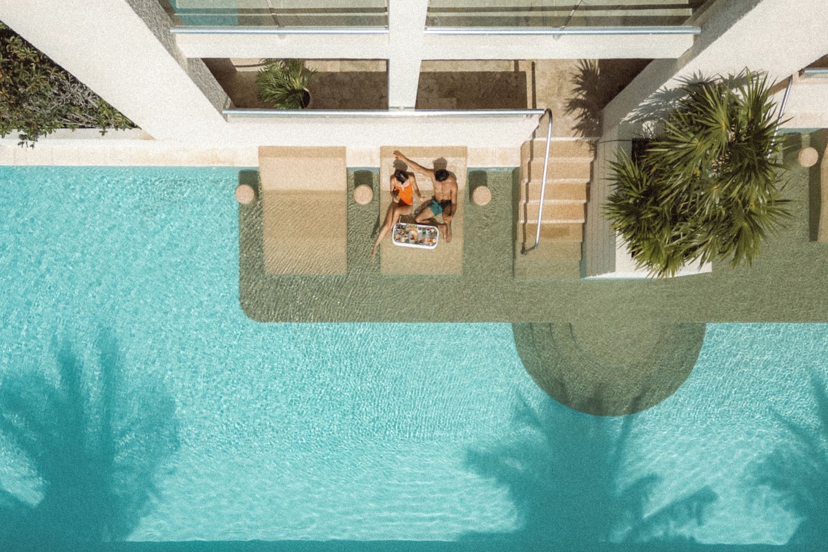An aerial view of a swimming pool with two people relaxing on the edge, surrounded by a modern building with large windows and a palm tree.