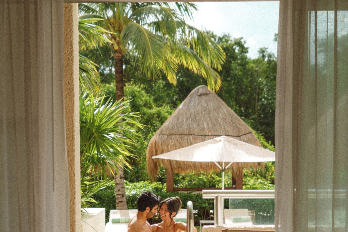A couple by a pool with a thatched umbrella, palm trees, and a view of the outdoors.