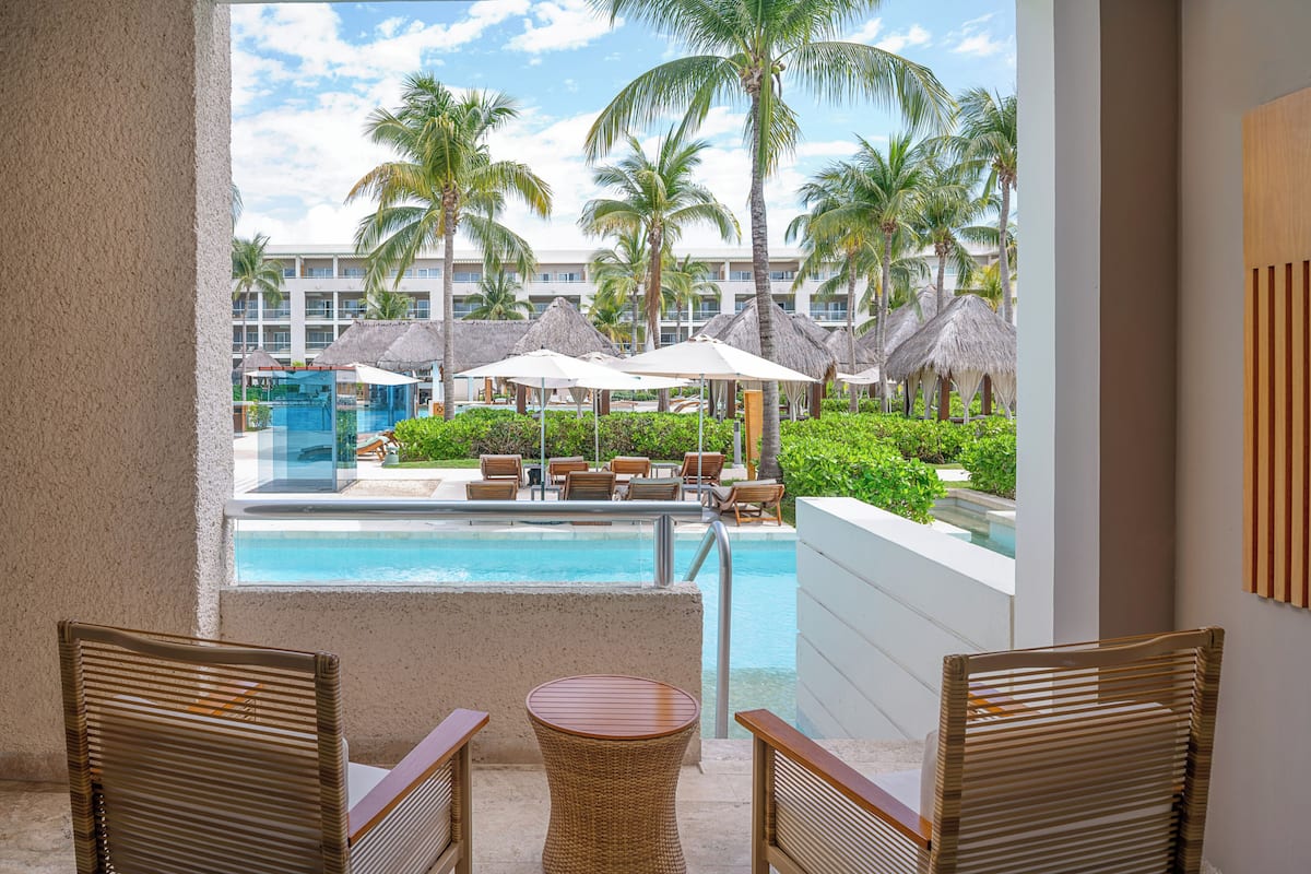 A balcony with wicker chairs and a small table, overlooking a pool area with umbrellas and palm trees.