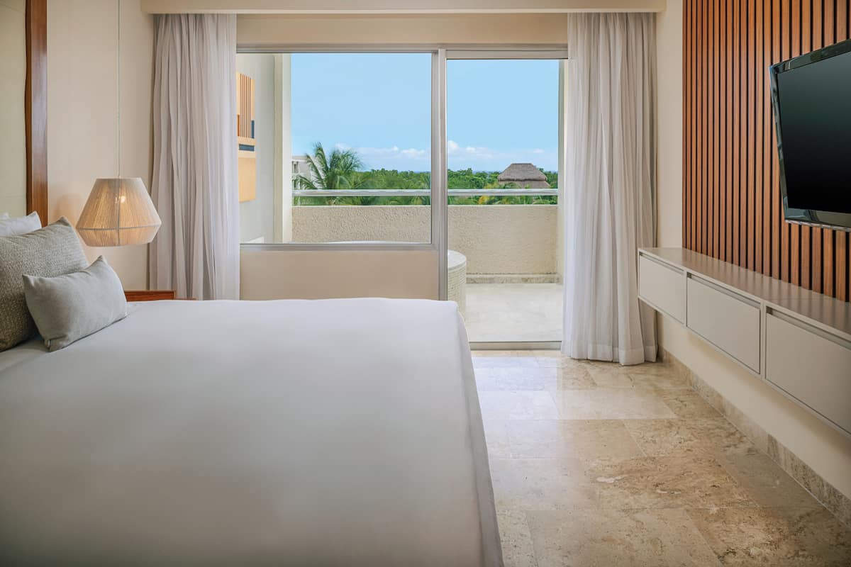 A hotel room with a bed, a TV, a balcony with a view of greenery and a building, and a tiled floor.