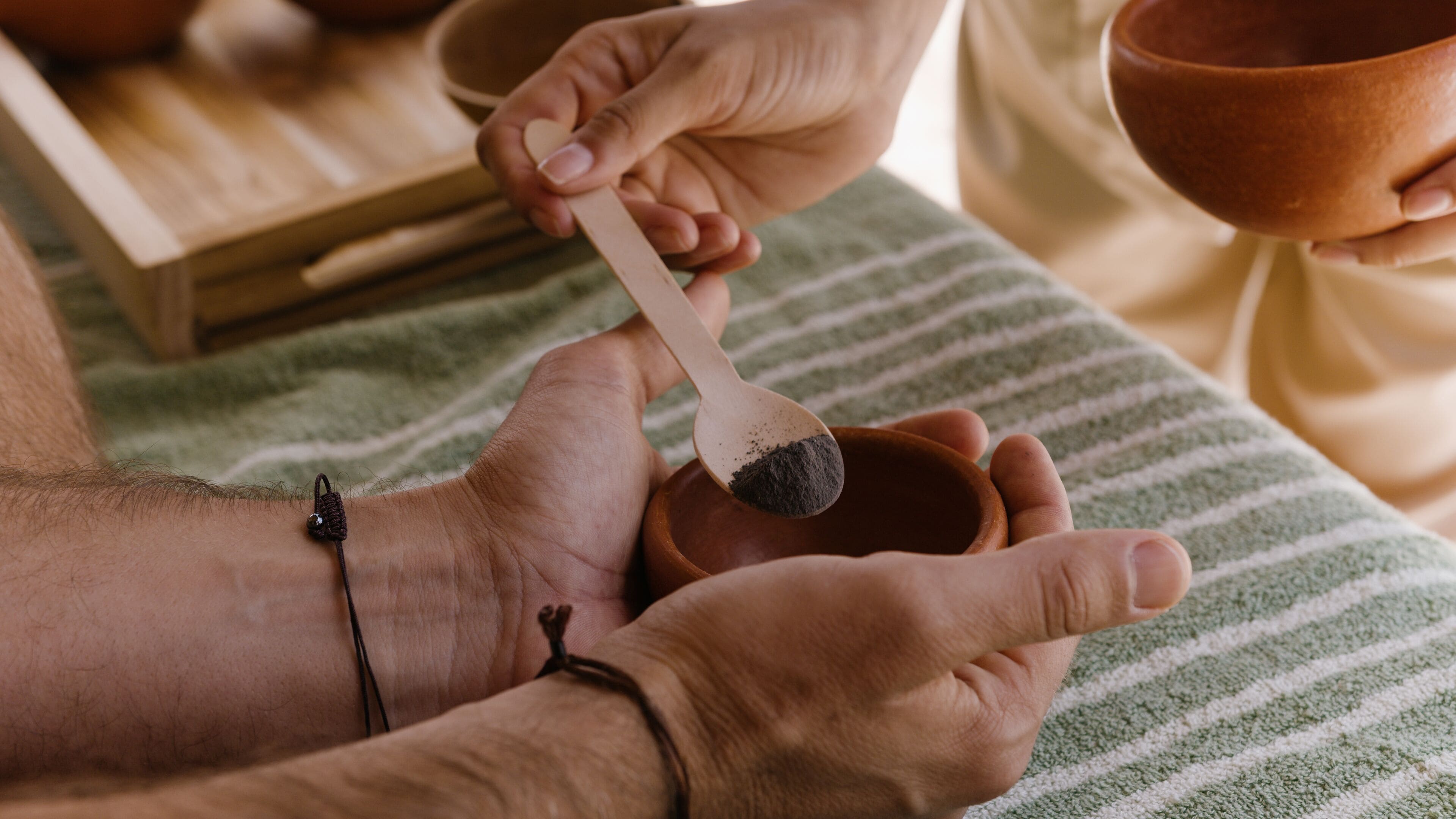 A person holding a wooden spoon with a dark powder, possibly for a spa treatment, with a wooden bowl nearby.