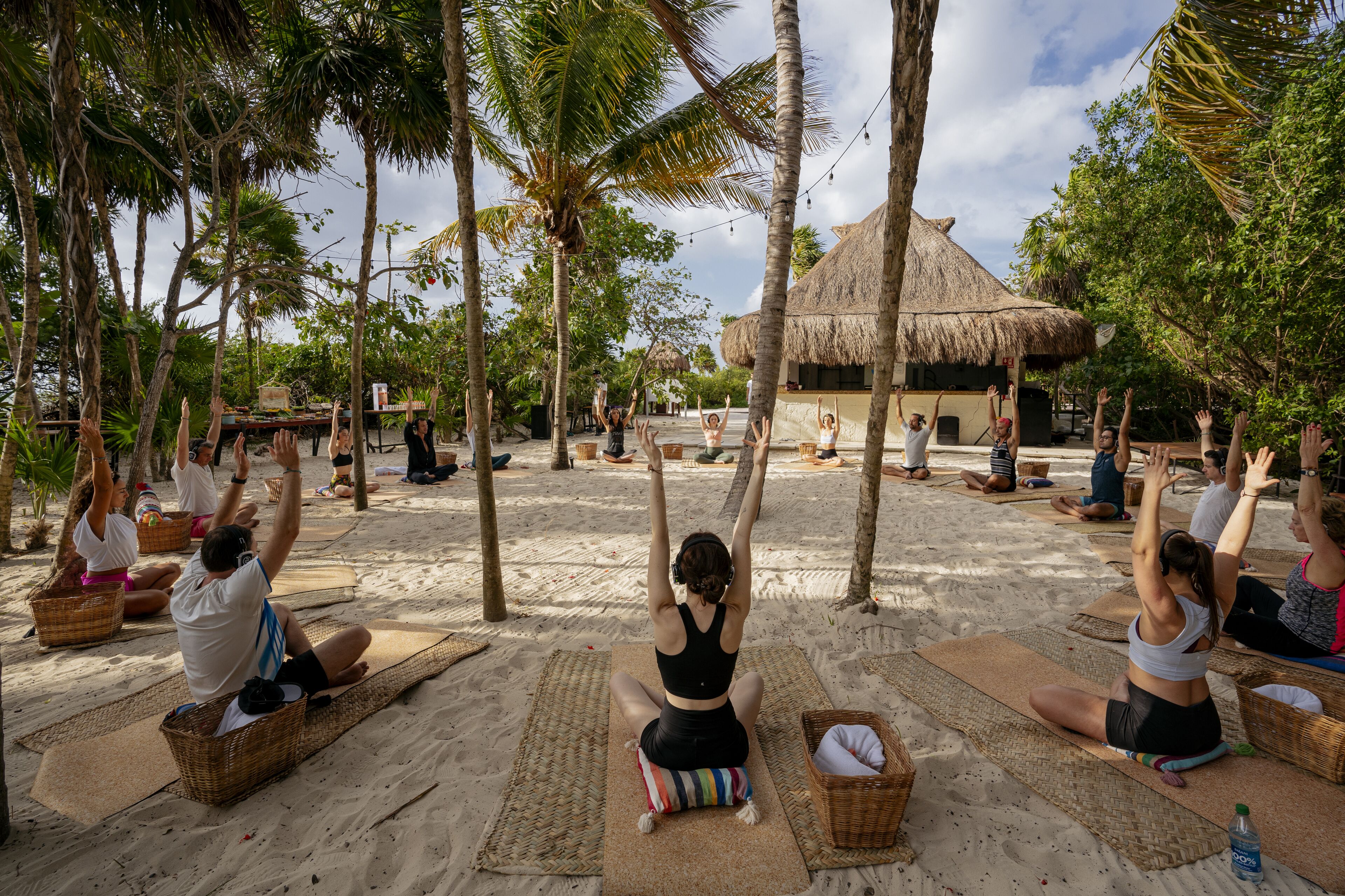 A group of people performing yoga on mats in a sandy area with trees and a thatched-roof structure in the background.