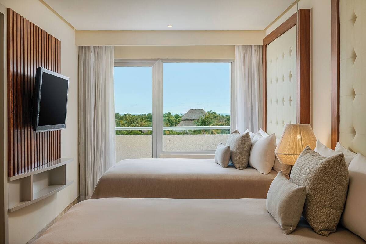 A hotel room with two beds, a TV, a lamp, and a view of the ocean.
