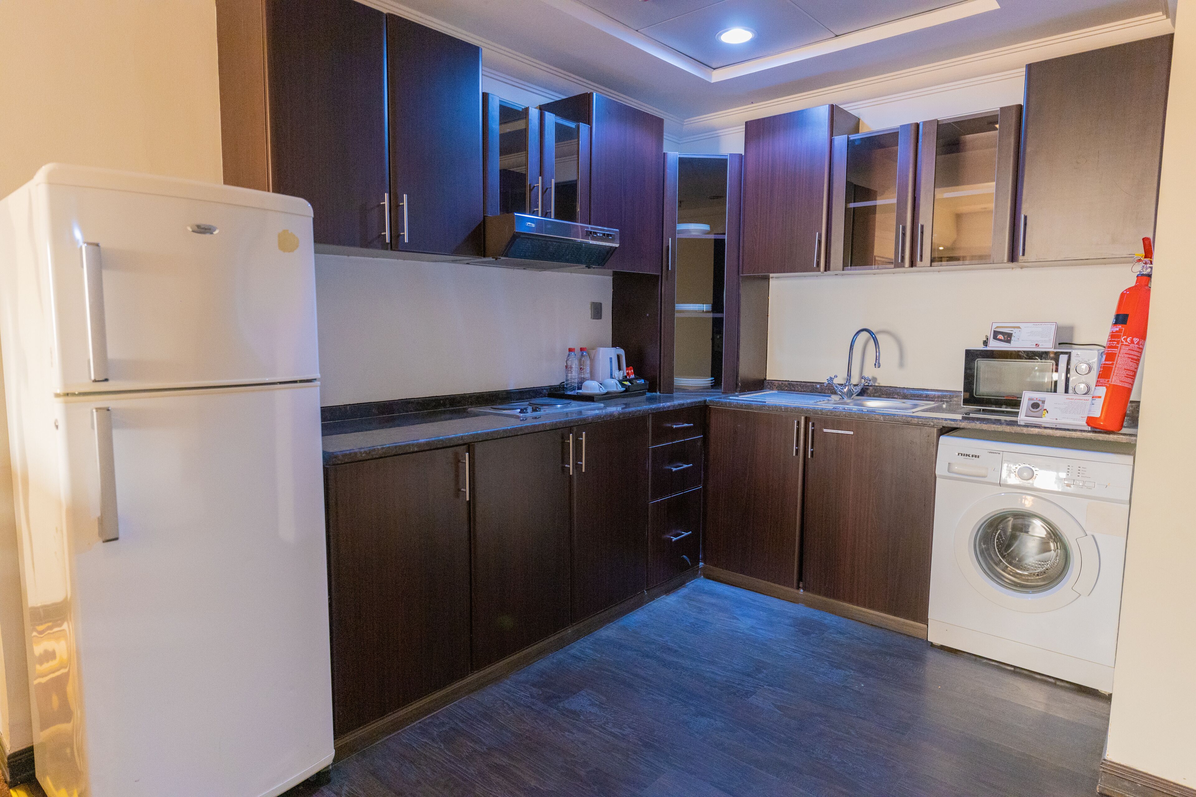 Una cocina compacta con armarios de madera oscura, un refrigerador blanco, un microondas y una lavadora.
