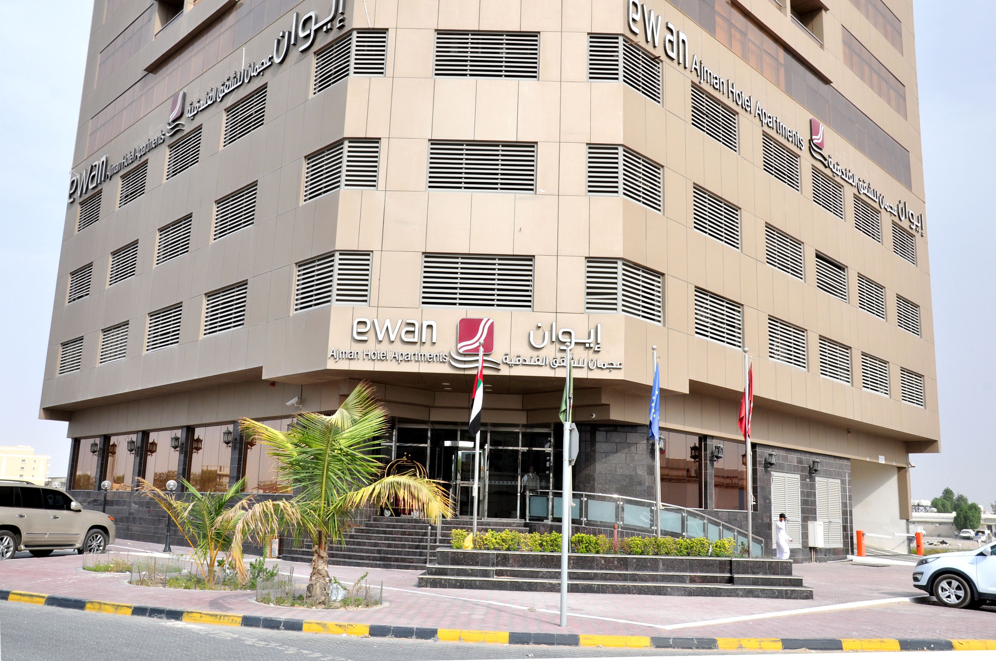 A modern building with the name 'Ewan Hotel Apartments' prominently displayed. It features a multi-story structure with a flat facade, several windows, and a set of stairs leading to the entrance.