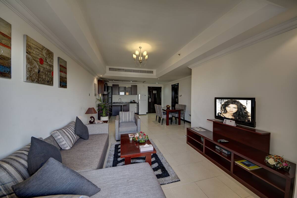 A modern living room with a sofa, coffee table, TV, and a dining area in the background.