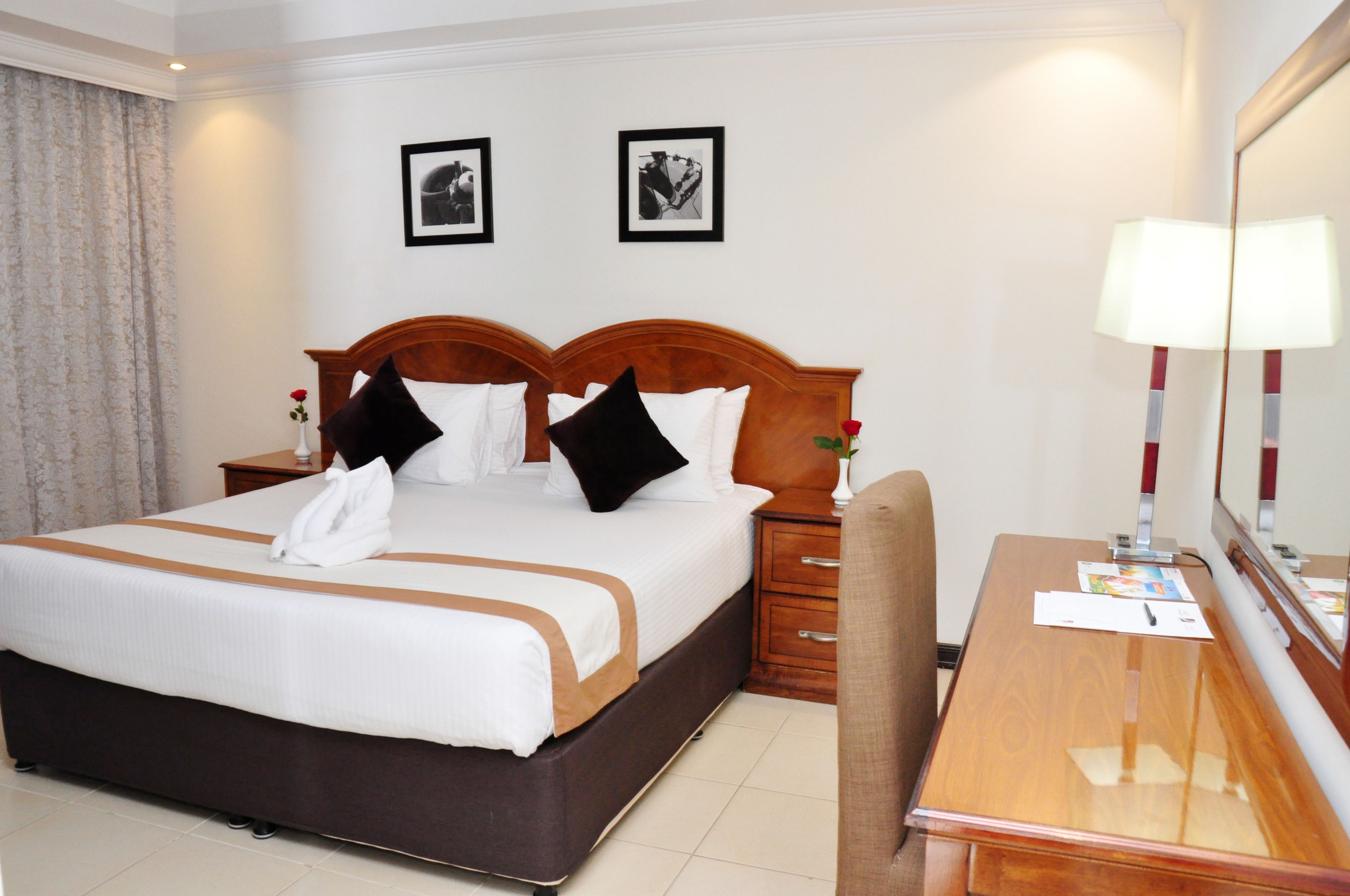 A hotel room with a large bed, a wooden headboard, a bedside table with a lamp, a desk with a mirror, and two framed pictures on the wall.