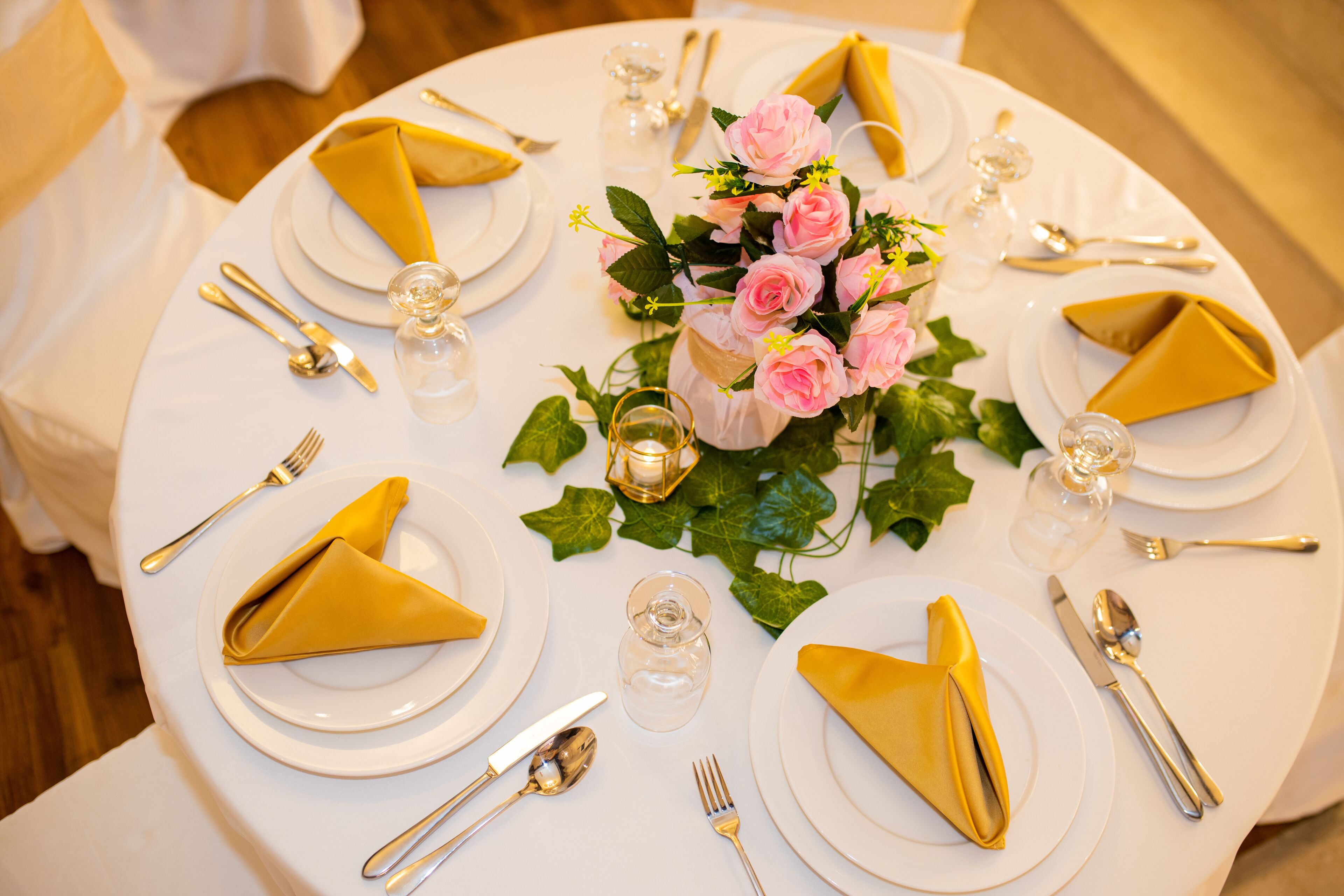 Una mesa redonda dispuesta para un evento formal, con mantel blanco, servilletas doradas y un centro de mesa con rosas rosadas.