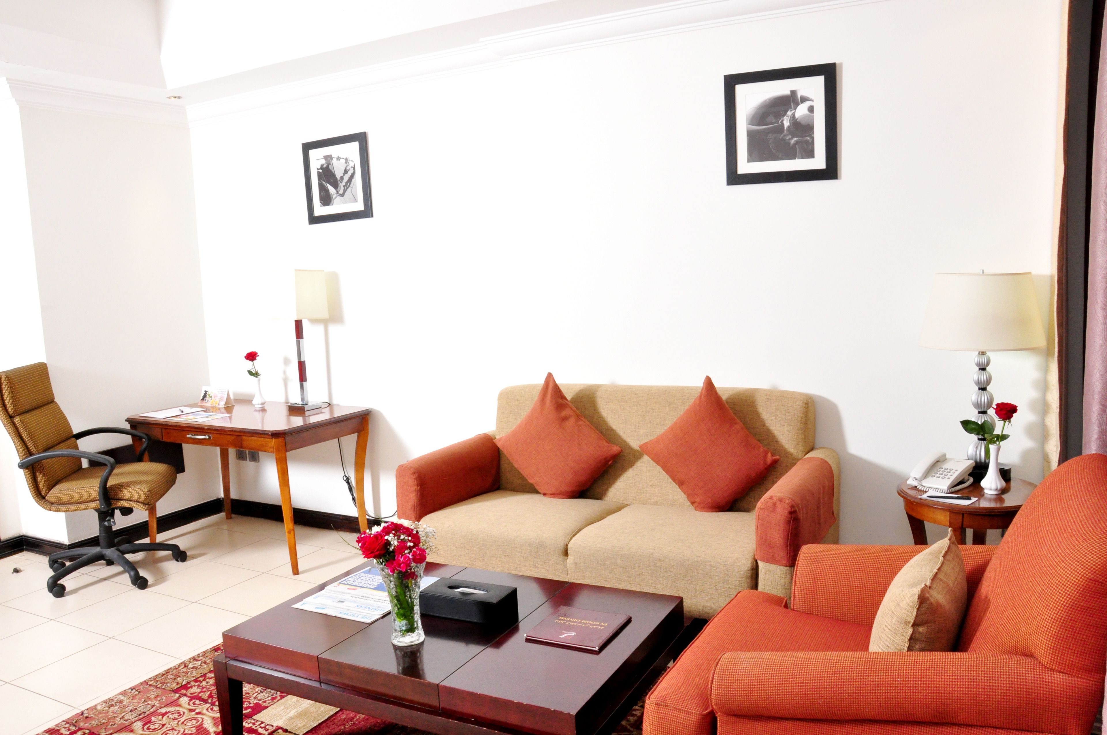 A living room with a beige sofa, two orange armchairs, a wooden coffee table, a desk with a lamp, and two framed pictures on the wall.