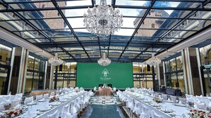 A banquet hall with a long table set for a formal event, a green screen with "The Chateau" logo, and a large chandelier.