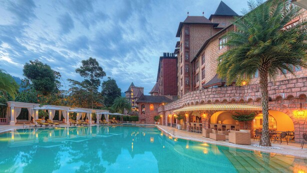 A large swimming pool with lounge chairs and umbrellas, surrounded by a stone building with arched windows and a tower.