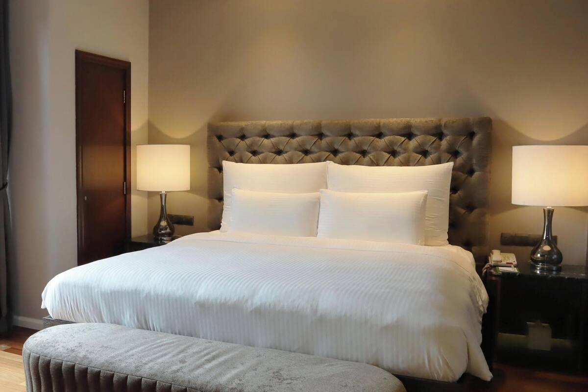 Une chambre d’hôtel avec un grand lit, deux tables de chevet avec des lampes, une porte en bois et un petit pouf.