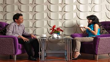 Two people seated on purple chairs in a modern room with a patterned wall and a small table with a white cup and a vase with red flowers.