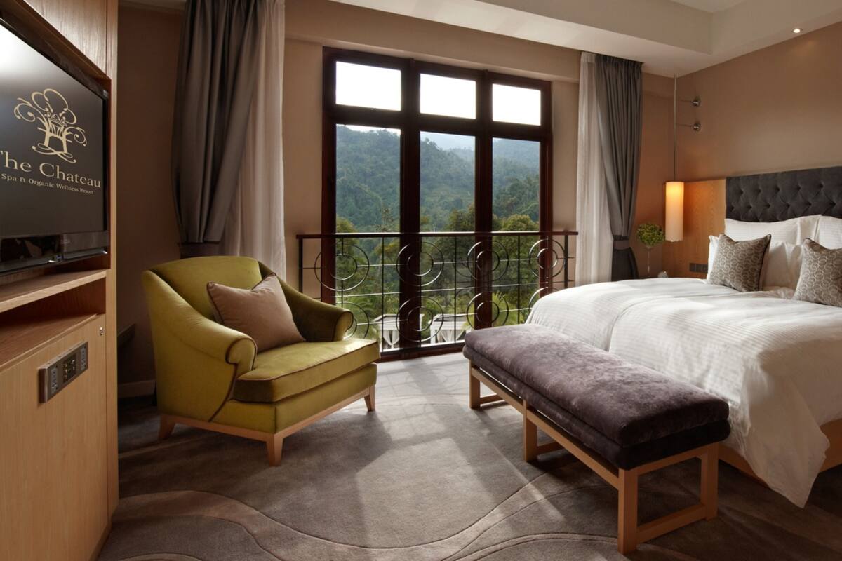 Une chambre d’hôtel avec un grand lit, un fauteuil vert, un banc, une télévision et un balcon offrant une vue panoramique.