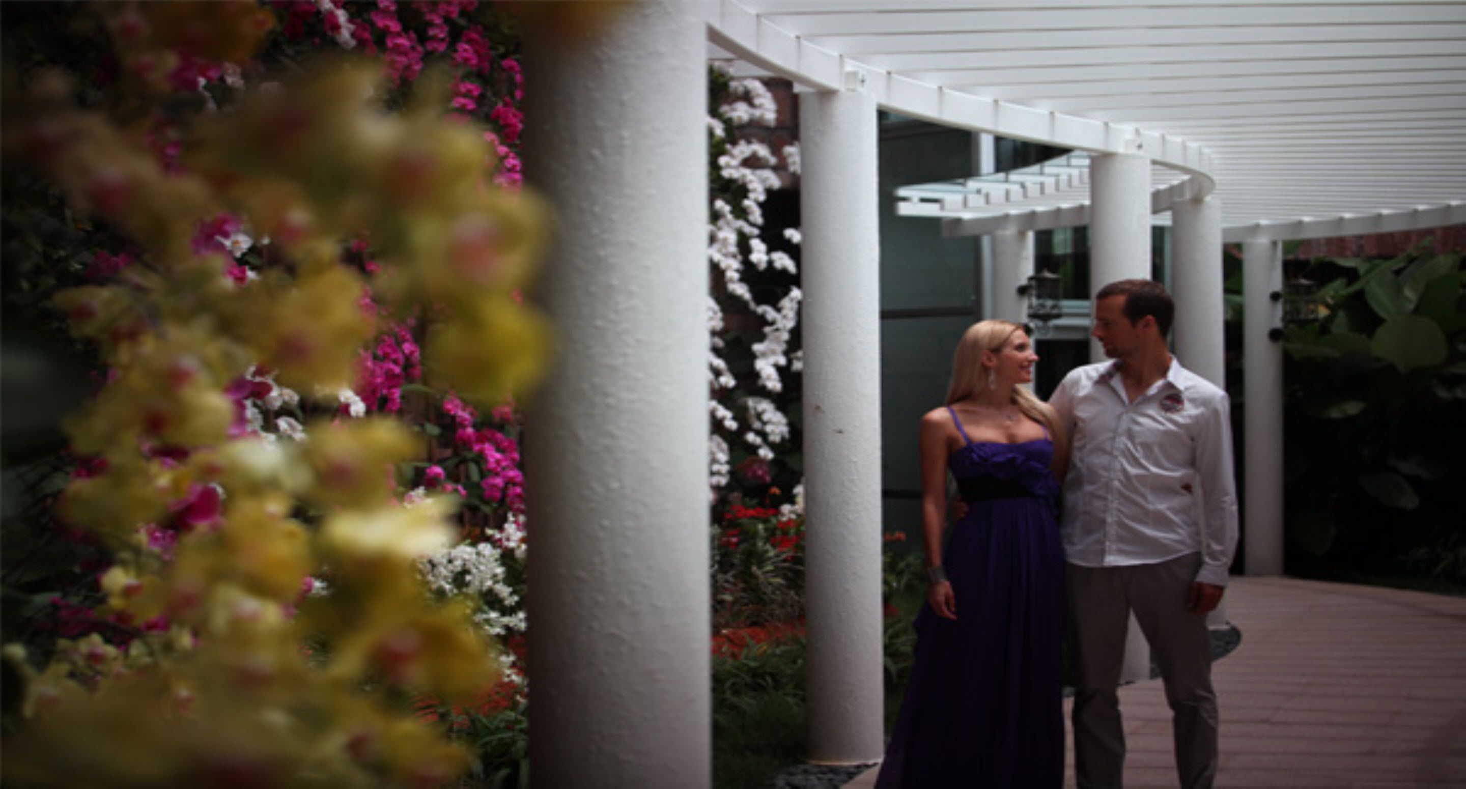 Una pareja está de pie en un espacio al aire libre cubierto, con columnas blancas y arreglos florales.