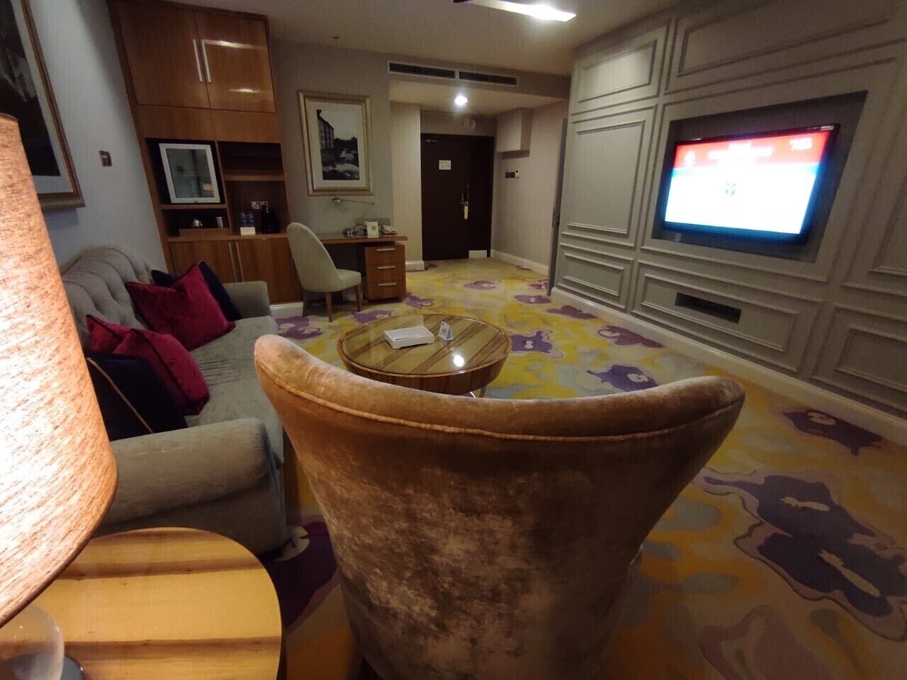 A living room with a sofa, armchair, and a coffee table. There is a television on the wall and a door in the background.