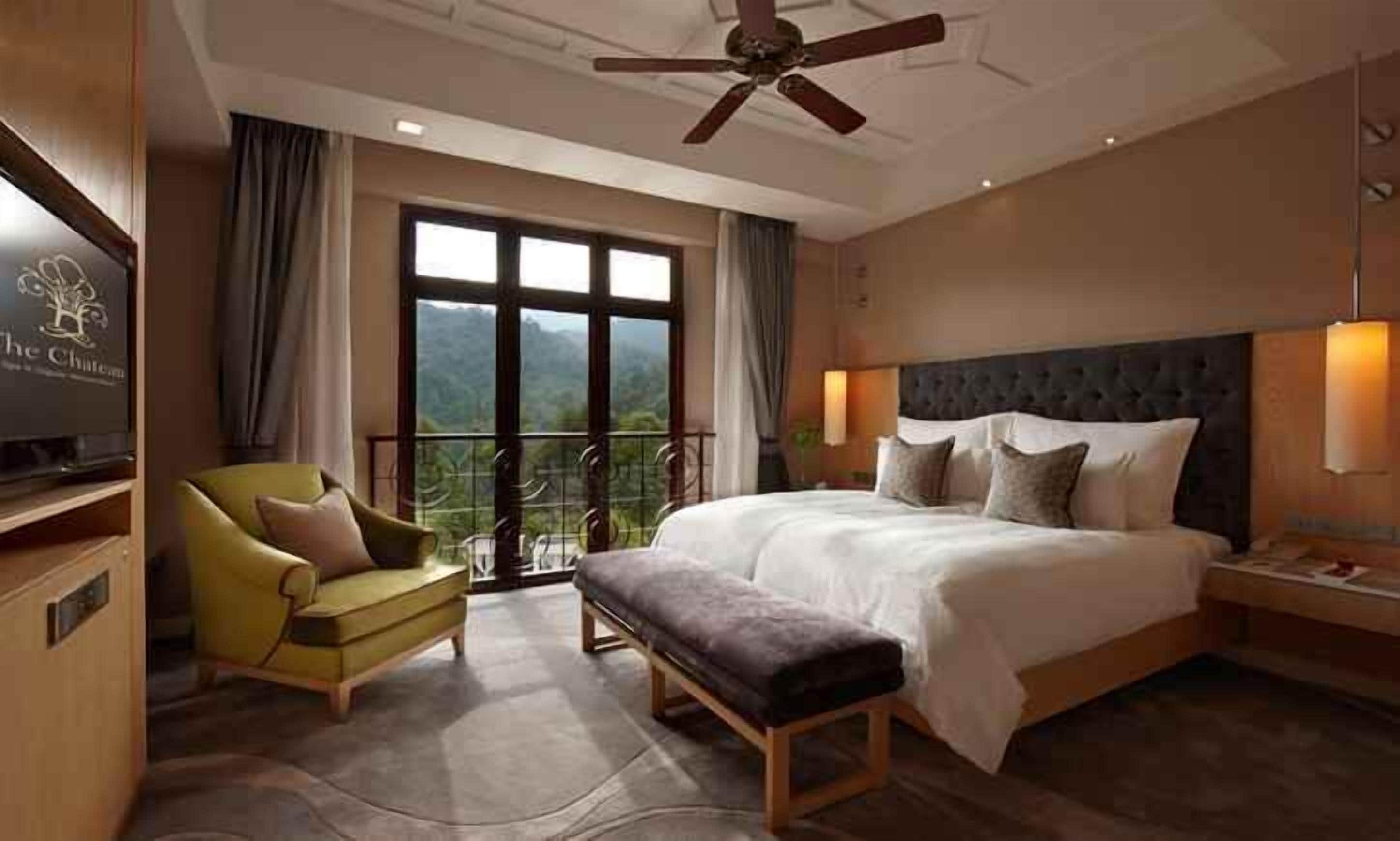 Habitación de hotel con una cama grande, un sofá, una banca, un televisor y un balcón con vistas.