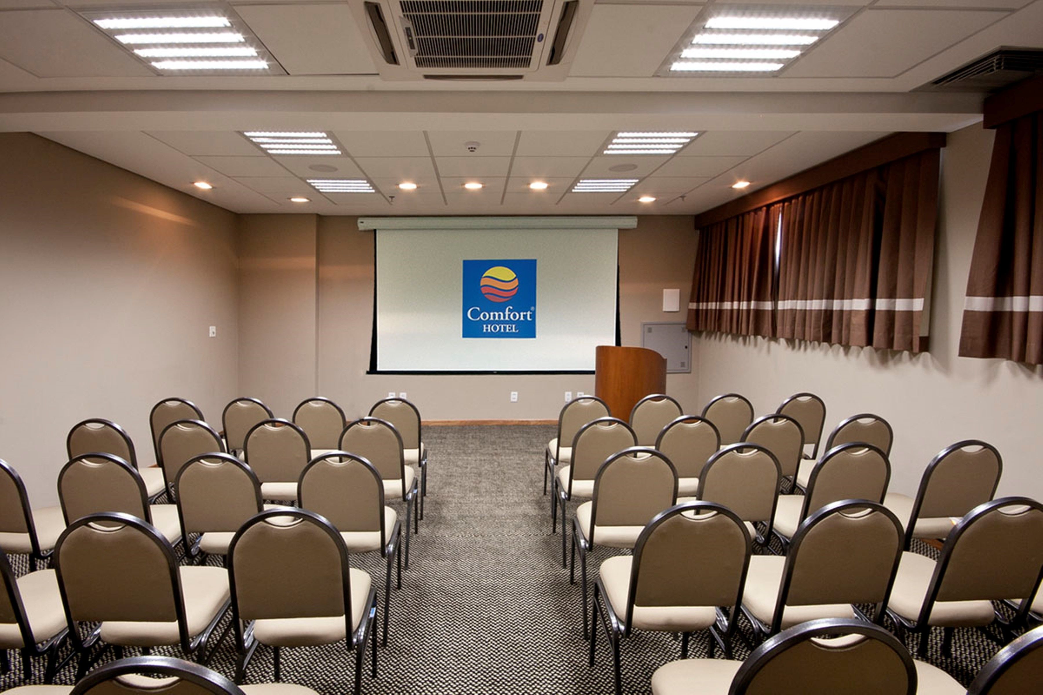 Een vergaderzaal met rijen stoelen gericht op een projectiescherm waarop het logo van Comfort Hotel staat.