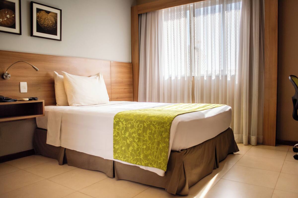 A hotel room with a bed, a bedside table, a lamp, a window with sheer curtains, and a wall-mounted light switch.