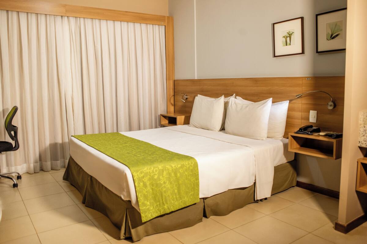 A hotel room with a large bed, a wooden headboard, a bedside table with a lamp, a chair, and a window with curtains.