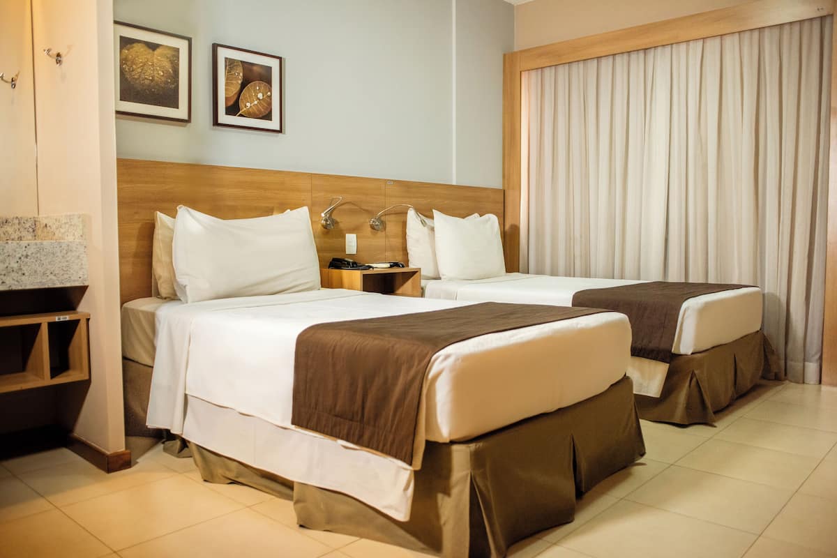 A hotel room with two beds, a wooden headboard, a nightstand, and a large window with curtains.