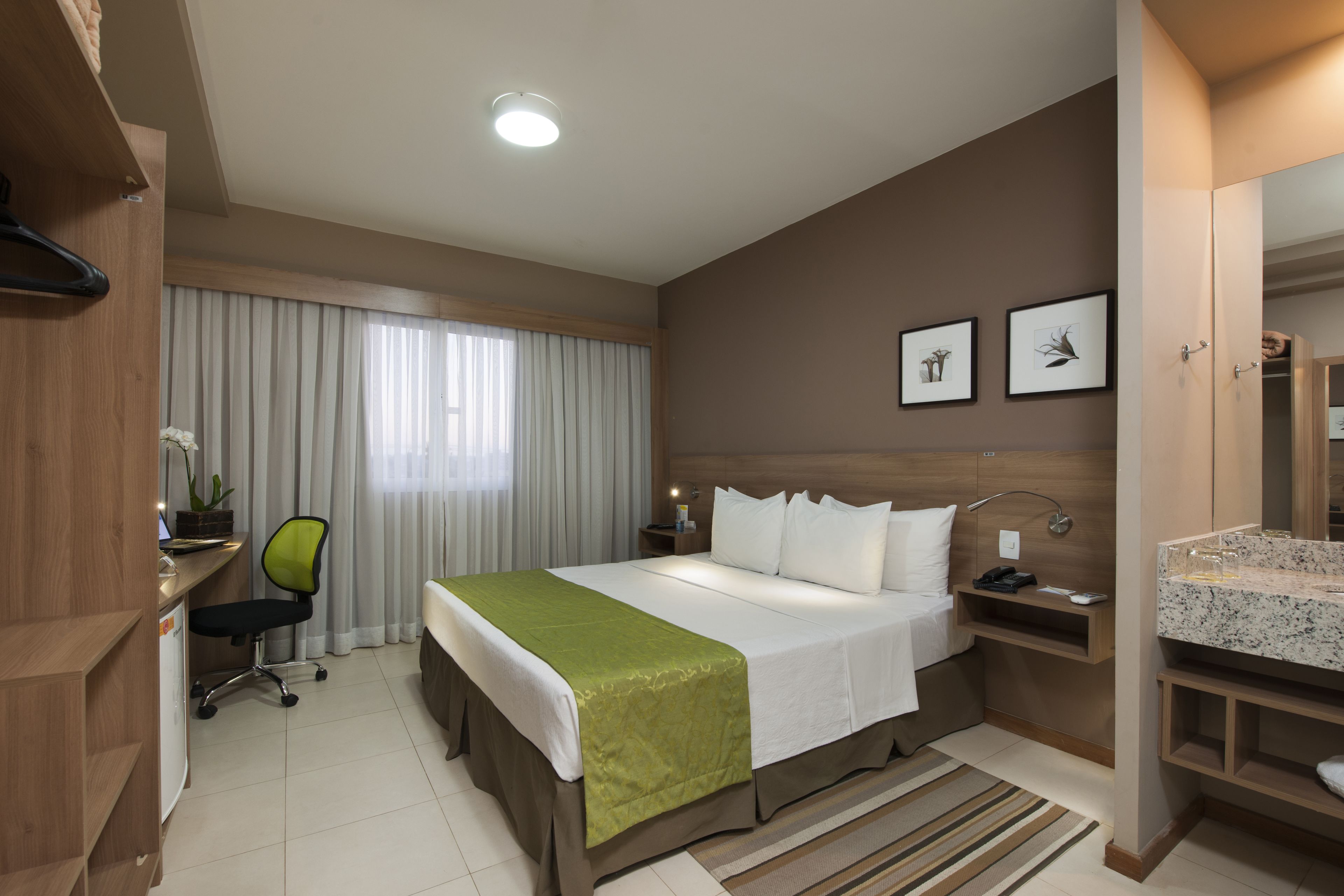 Habitación de hotel con cama, escritorio, silla, mesita de noche y baño con lavamanos y espejo.